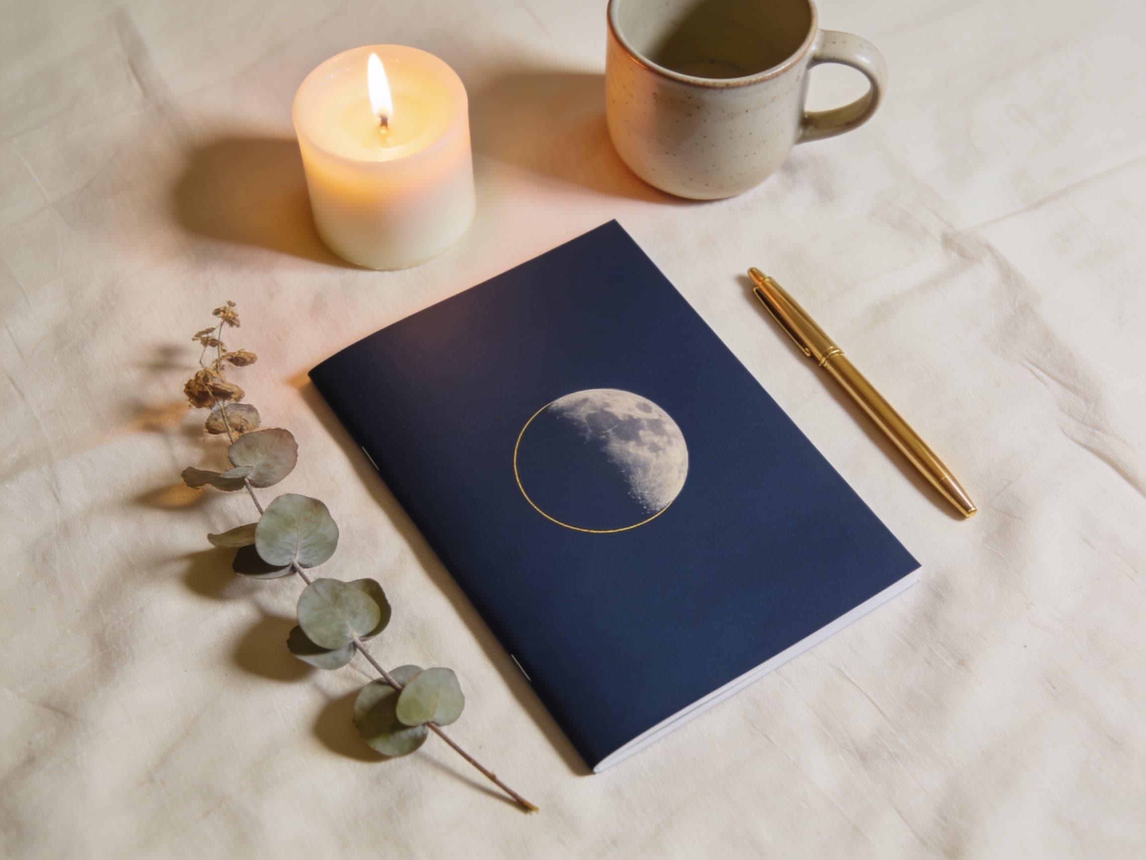 The Full Moon Money Date ritual guide — an indigo booklet stamped with a gold moon, beside a lit candle, a mug, and eucalyptus on linen.
